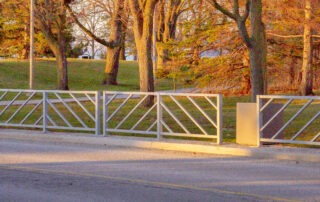 A metal gate with diagonal patterns stands across a road. Behind it, a grassy area dotted with bare and evergreen trees reflects the changing seasons under a clear sky. Sunlight casts long shadows on the grass, suggesting either sunrise or sunset.