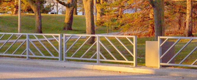 A metal gate with diagonal patterns stands across a road. Behind it, a grassy area dotted with bare and evergreen trees reflects the changing seasons under a clear sky. Sunlight casts long shadows on the grass, suggesting either sunrise or sunset.