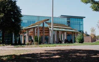 A modern building with large glass windows stands behind an inspiring pavilion, serving as an outdoor gathering space with a wooden roof structure. People mingle beneath it, embraced by trees and greenery in the foreground, under a clear blue sky.