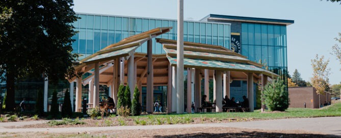 A modern building with large glass windows stands behind an inspiring pavilion, serving as an outdoor gathering space with a wooden roof structure. People mingle beneath it, embraced by trees and greenery in the foreground, under a clear blue sky.