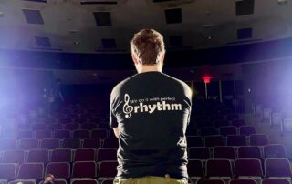 A person stands on stage in a dimly lit theater, their black t-shirt proclaiming "We do it with perfect rhythm," embodying artistic expression. Two spotlights shine down, illuminating the empty seats and hinting at comedic possibilities yet to unfold.