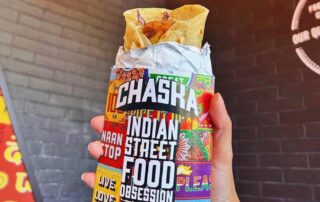 A hand holds a wrapped Indian street food roll labeled "CHASKA," brimming with colorful graphics and phrases like "NAAN STOP" and "LIVE LOVE." Against an open brick wall backdrop with partial signage, this captures the vibrant essence of Chaska.