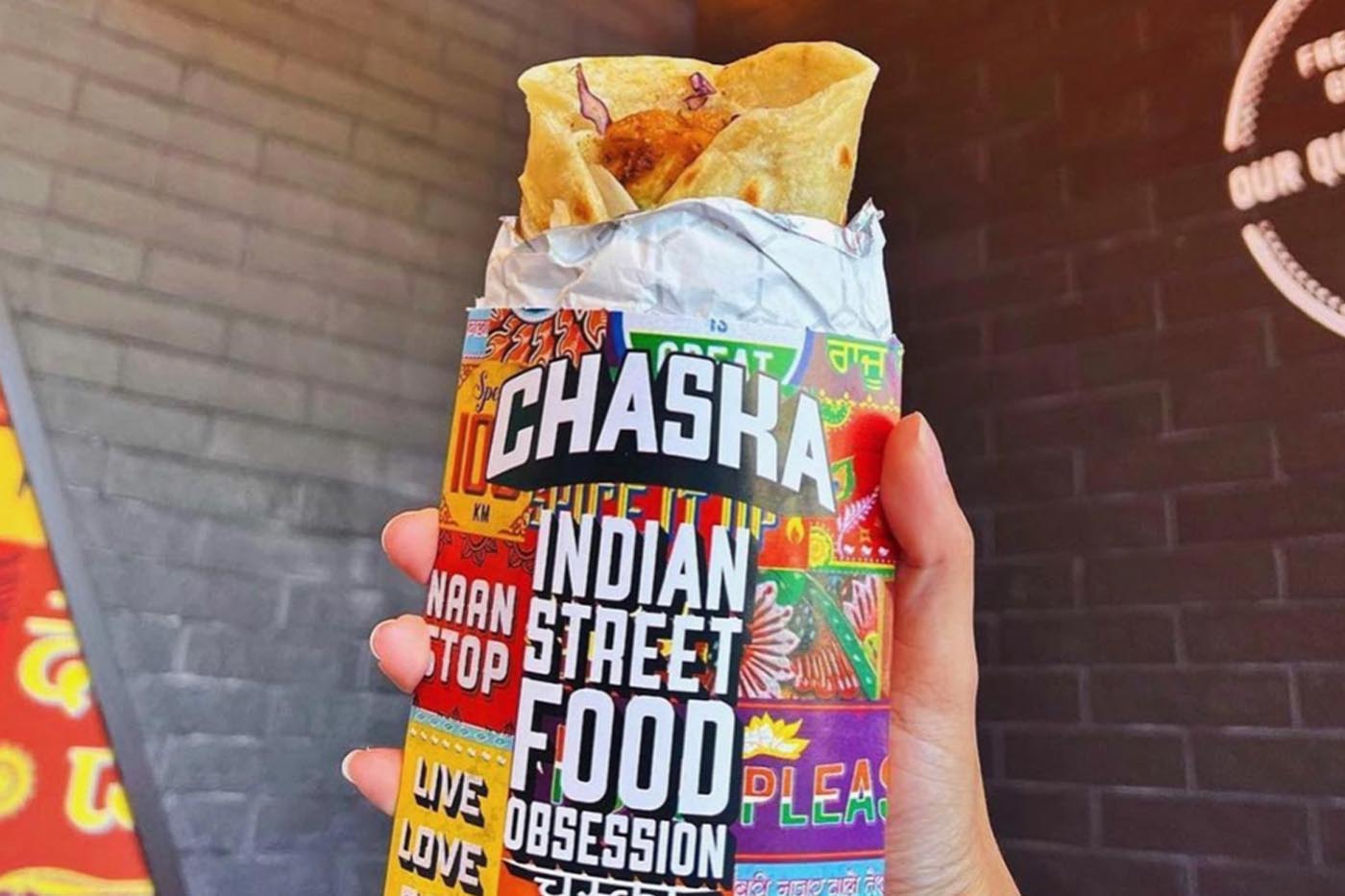 A hand holds a wrapped Indian street food roll labeled "CHASKA," brimming with colorful graphics and phrases like "NAAN STOP" and "LIVE LOVE." Against an open brick wall backdrop with partial signage, this captures the vibrant essence of Chaska.