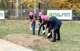 Four people in pink hard hats participate in a groundbreaking ceremony for a new mathematics building, using shovels to dig into a small mound of earth. They stand outdoors in a grassy area, surrounded by autumn leaves and construction banners in the background.