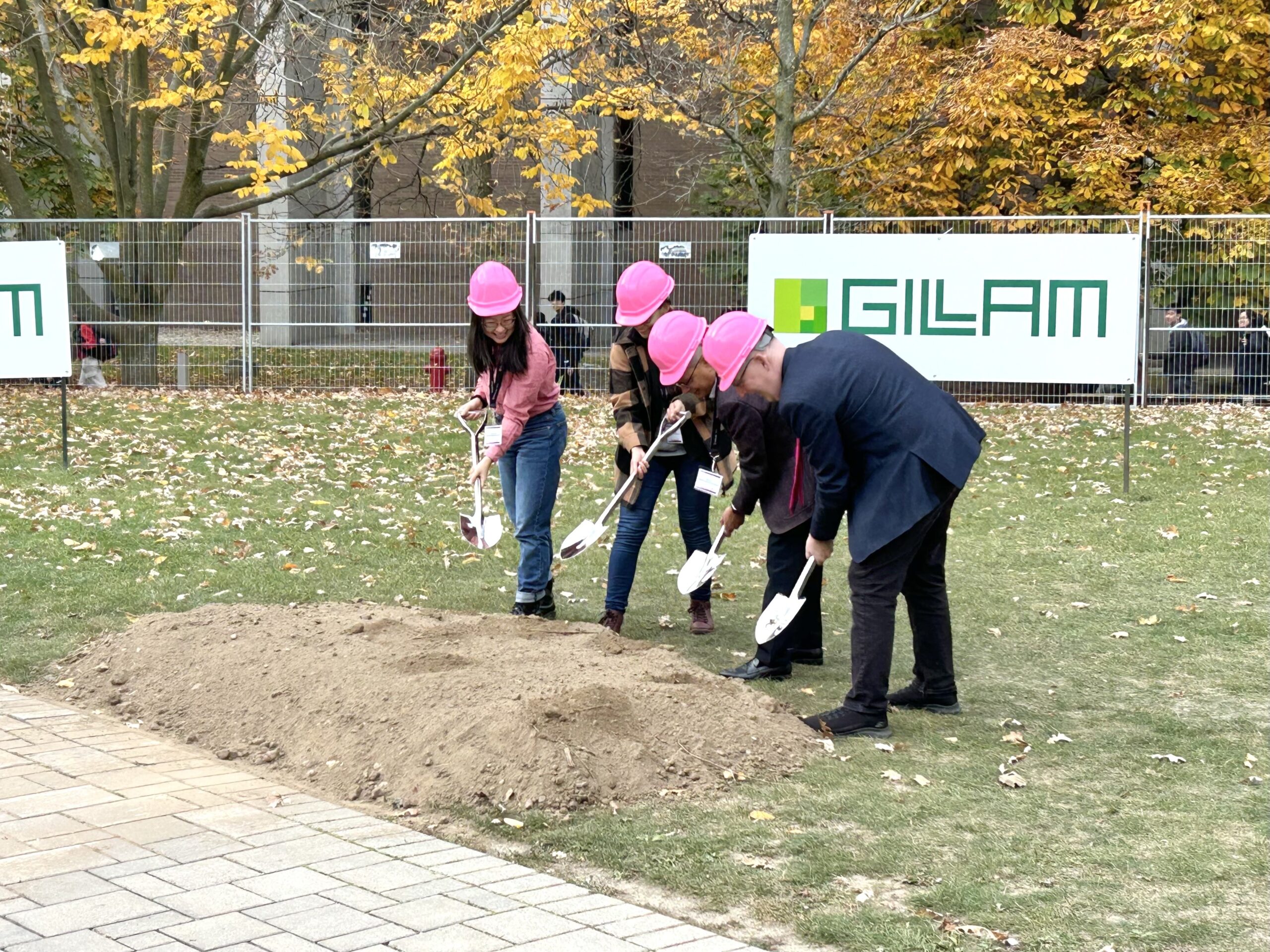 A group of people in pink hard hats are celebrating the groundbreaking ceremony for a new mathematics building. They enthusiastically dig into a small mound of dirt with their shovels, while trees with autumn leaves provide a scenic backdrop and signs adorn the fence behind them.