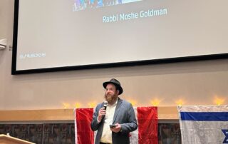 A man stands at a podium, speaking into a microphone. Behind him is a screen displaying a group photo and text "Rabbi Moshe Goldman." Canadian and Israeli flags are draped below the screen. Flowers are arranged on the podium as he addresses those remembering one year later.