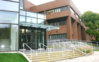 A modern brick and glass building graces the diverse campus, featuring an entrance with glass doors and a canopy. Multiple sets of stairs and ramps with railings lead to the entrance, while a tree stands tall on the right side, offering a welcoming touch.