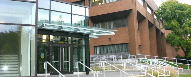 A modern brick and glass building graces the diverse campus, featuring an entrance with glass doors and a canopy. Multiple sets of stairs and ramps with railings lead to the entrance, while a tree stands tall on the right side, offering a welcoming touch.