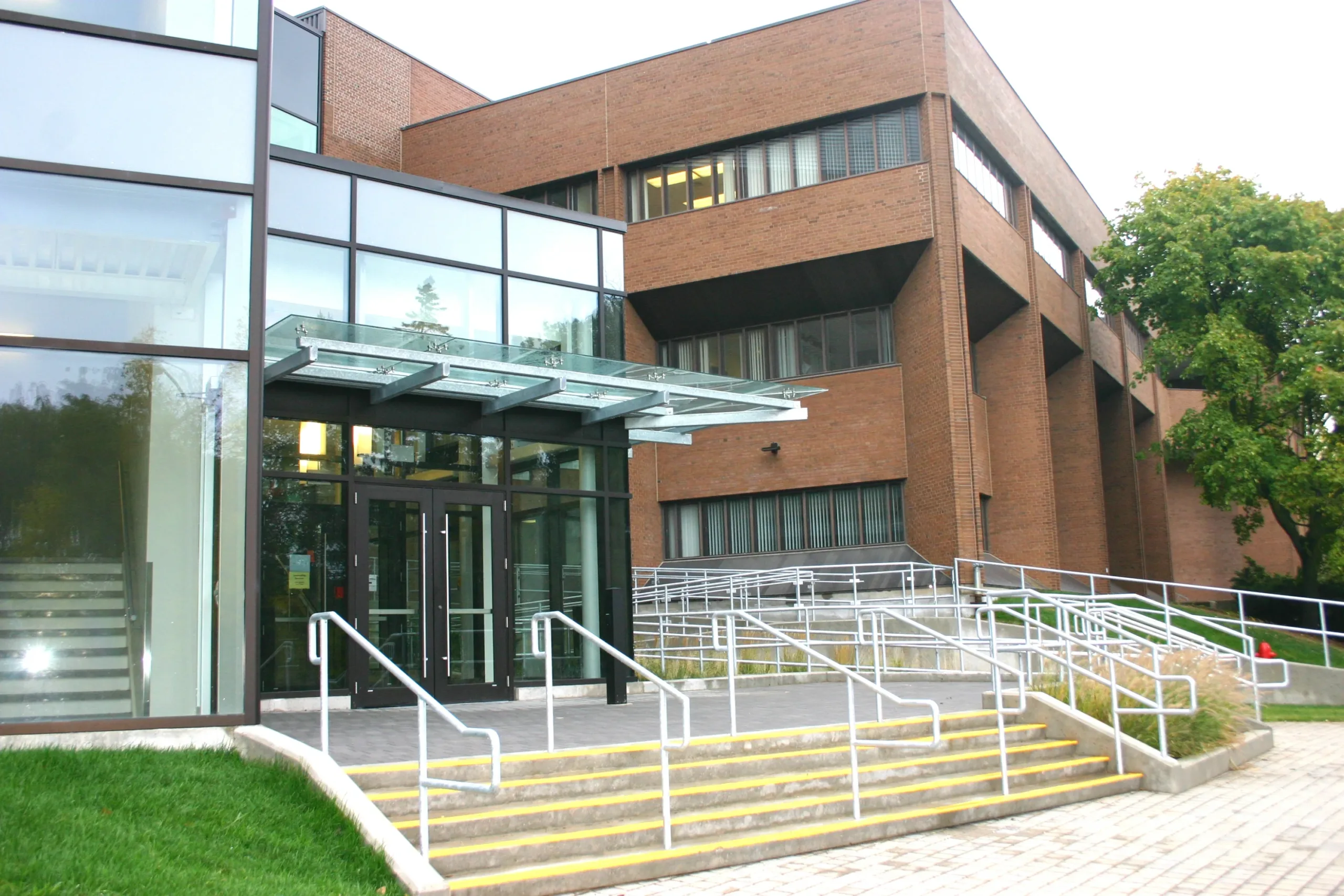 A modern brick and glass building graces the diverse campus, featuring an entrance with glass doors and a canopy. Multiple sets of stairs and ramps with railings lead to the entrance, while a tree stands tall on the right side, offering a welcoming touch.