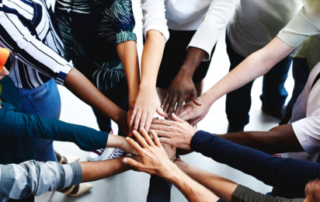 A diverse group of people from UW stands in a circle, their hands reaching toward the center and stacked on top of each other, symbolizing teamwork and unity. Their varied clothing styles and colors showcase the inclusive spirit of an EDI-focused research hub.