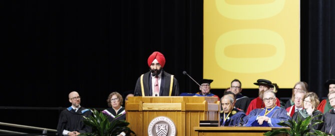 Jagdeep Singh Bachher, donning a red turban, stands at the podium during the graduation ceremony. The UW chancellor is flanked by faculty in academic regalia, with a large yellow banner behind them. Lush plants add a touch of greenery to the celebratory stage.