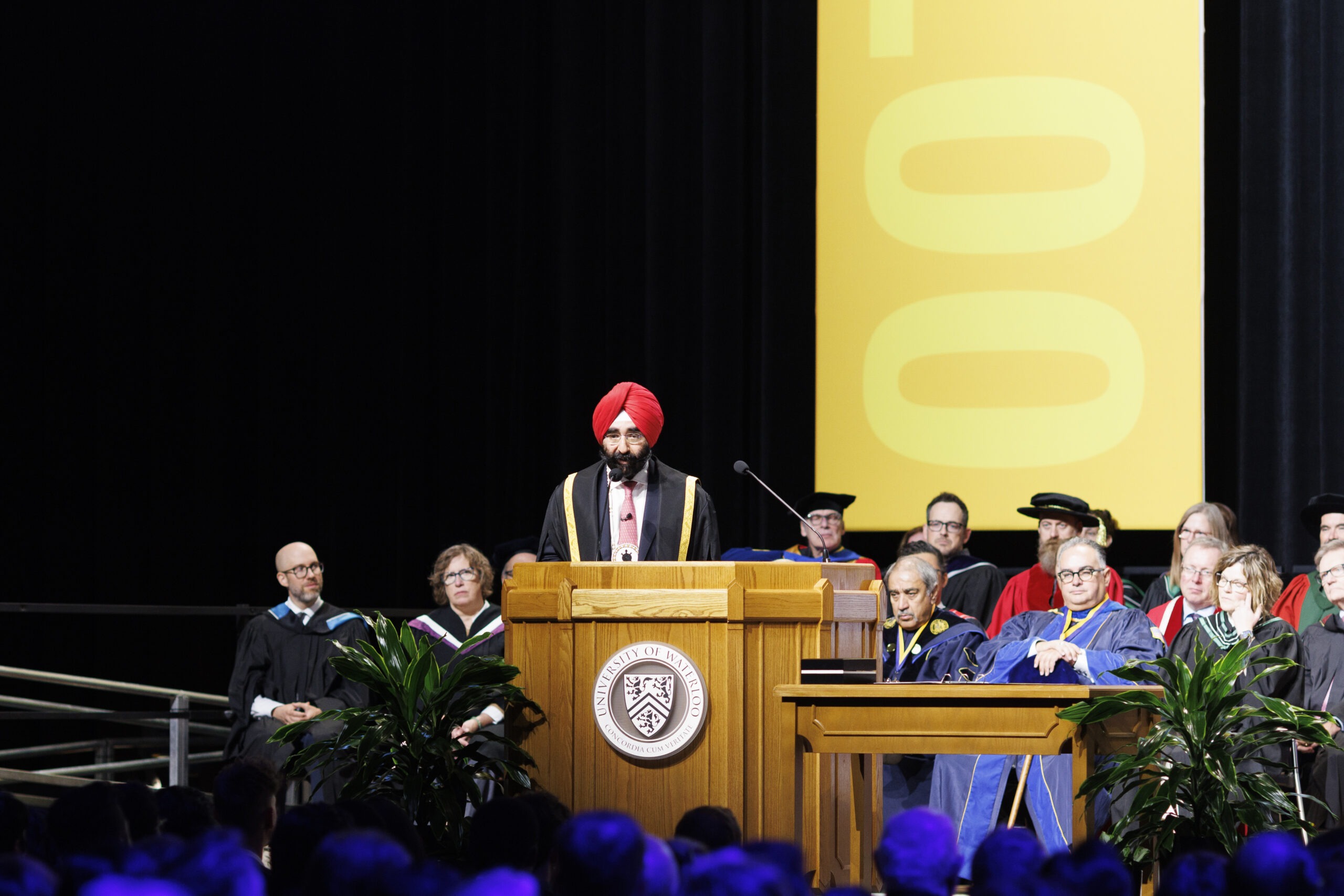 Jagdeep Singh Bachher, donning a red turban, stands at the podium during the graduation ceremony. The UW chancellor is flanked by faculty in academic regalia, with a large yellow banner behind them. Lush plants add a touch of greenery to the celebratory stage.