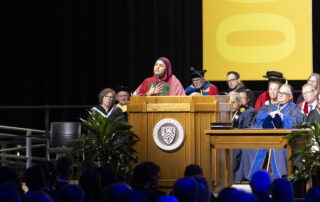 A person in a red outfit and headscarf speaks passionately at the podium during the graduation ceremony, sharing valuable graduate advice. Behind them, faculty members in academic regalia sit on stage, with a large yellow banner visible in the background.