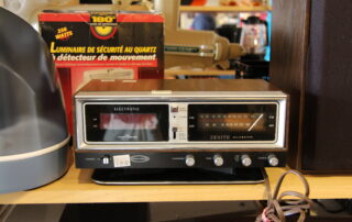 A vintage Zenith clock radio sits on a shelf with a brown casing and a digital display. To the left is a box of a motion detector light, and cables are visible in the foreground.