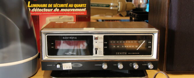 A vintage Zenith clock radio sits on a shelf with a brown casing and a digital display. To the left is a box of a motion detector light, and cables are visible in the foreground.