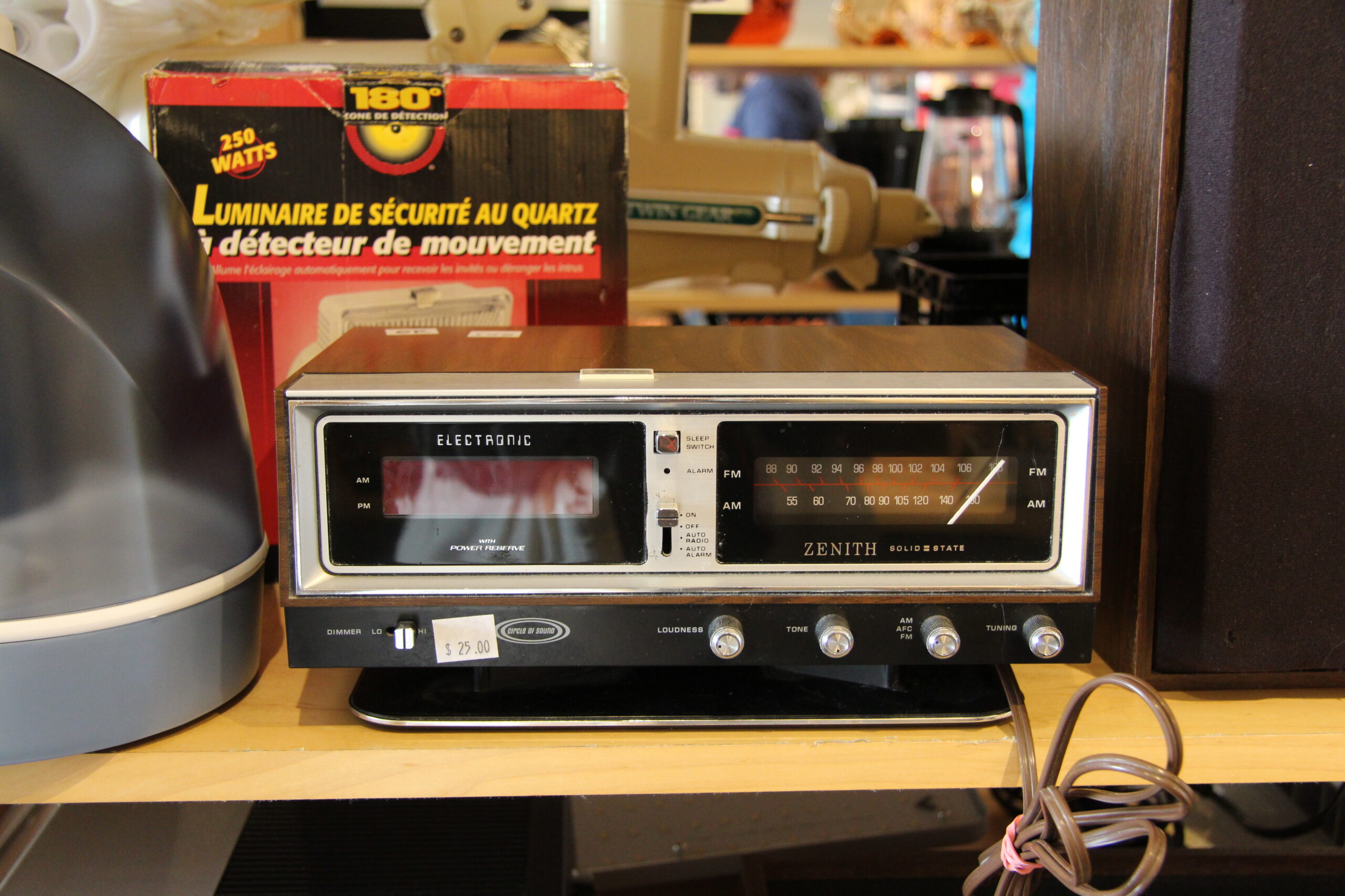 A vintage Zenith clock radio sits on a shelf with a brown casing and a digital display. To the left is a box of a motion detector light, and cables are visible in the foreground.