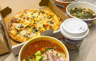 A variety of foods on a table includes a pizza in a box, two bowls of spicy Indian dishes with vegetables and onions, a cup of hot drink, and a smoothie topped with nuts.