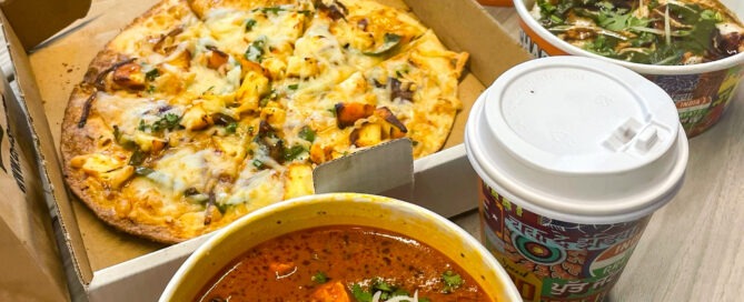 A variety of foods on a table includes a pizza in a box, two bowls of spicy Indian dishes with vegetables and onions, a cup of hot drink, and a smoothie topped with nuts.