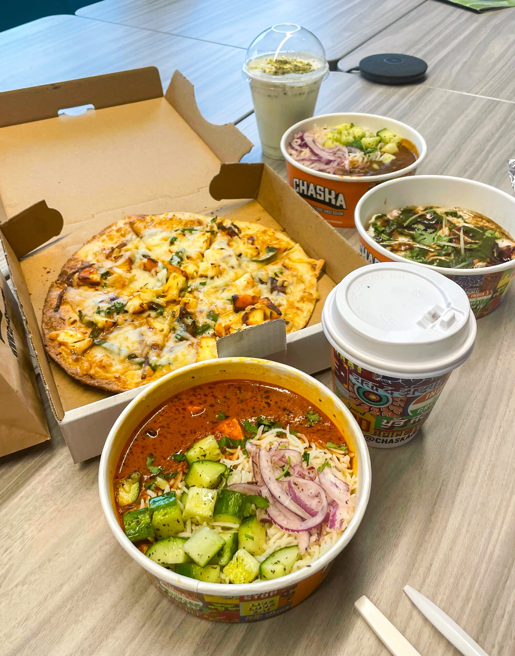 A variety of foods on a table includes a pizza in a box, two bowls of spicy Indian dishes with vegetables and onions, a cup of hot drink, and a smoothie topped with nuts.