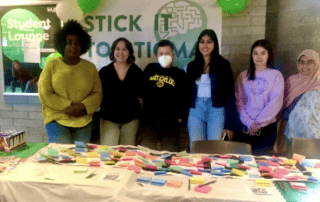 A group of six people stands behind a table adorned with colorful sticky notes and QR codes, creating a vibrant canvas of conversation. A "Stick It To Stigma" banner hangs proudly on the wall behind them, while green and white balloons add a festive touch to the scene.