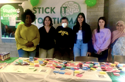 A group of six people stands behind a table adorned with colorful sticky notes and QR codes, creating a vibrant canvas of conversation. A "Stick It To Stigma" banner hangs proudly on the wall behind them, while green and white balloons add a festive touch to the scene.