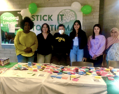 A group of six people stands behind a table adorned with colorful sticky notes and QR codes, creating a vibrant canvas of conversation. A "Stick It To Stigma" banner hangs proudly on the wall behind them, while green and white balloons add a festive touch to the scene.