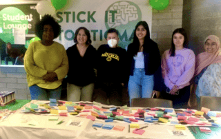 A group of six people stands behind a table covered with colorful sticky notes, engaging in conversation. They are at an event with "Stick it to Stigma" displayed on a banner. Green and white balloons decorate the area, creating a lively canvas of colors and ideas.