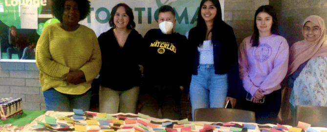 A group of six people stands behind a table covered with colorful sticky notes, engaging in conversation. They are at an event with "Stick it to Stigma" displayed on a banner. Green and white balloons decorate the area, creating a lively canvas of colors and ideas.