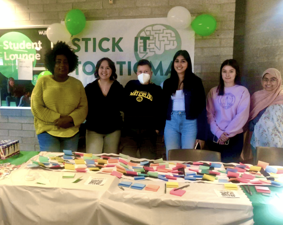 A group of six people stands behind a table covered with colorful sticky notes, engaging in conversation. They are at an event with "Stick it to Stigma" displayed on a banner. Green and white balloons decorate the area, creating a lively canvas of colors and ideas.