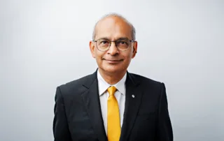 Elderly man with glasses wearing a black suit, white shirt, and yellow tie stands against a plain white background, smiling slightly at the camera. Could this poised gentleman be our future 2026 President? Perhaps he is drawing inspiration from leaders like Vivek Goel.