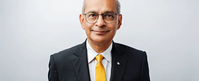 Elderly man with glasses wearing a black suit, white shirt, and yellow tie stands against a plain white background, smiling slightly at the camera. Could this poised gentleman be our future 2026 President? Perhaps he is drawing inspiration from leaders like Vivek Goel.