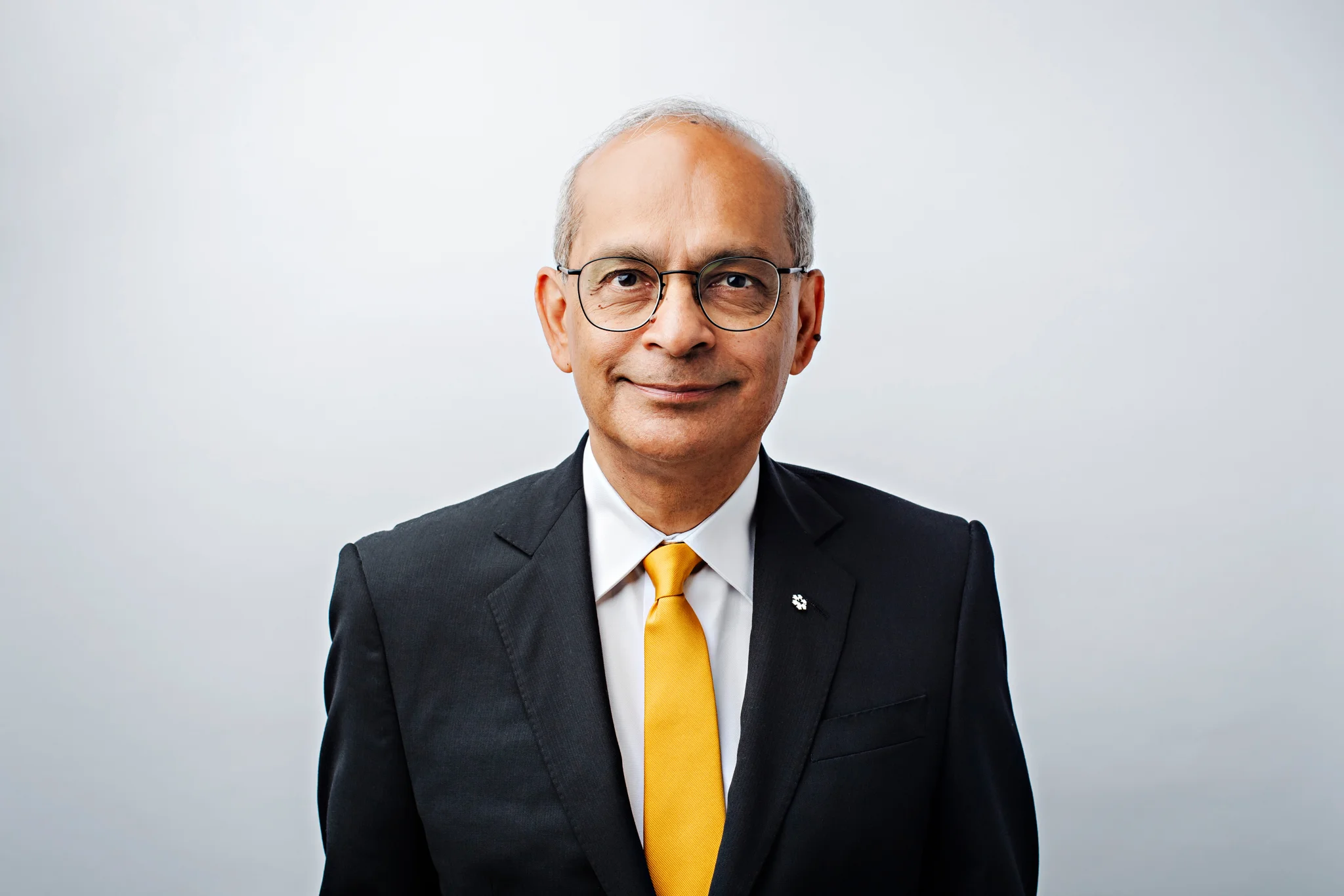 Elderly man with glasses wearing a black suit, white shirt, and yellow tie stands against a plain white background, smiling slightly at the camera. Could this poised gentleman be our future 2026 President? Perhaps he is drawing inspiration from leaders like Vivek Goel.