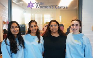 Four people stand smiling in front of a door labeled "WUSA Women's Centre," promoting equity in support services. Three are wearing matching light blue shirts, and one is donning a black shirt. The door features a purple logo highlighting the center's focus on menstrual wellness with silhouettes of several women.