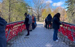 People stand on a cobblestone bridge tying red ribbons to the railing as part of a poignant bridge installation. The scene, set in a park with trees displaying autumn foliage under a clear blue sky, honors missing and murdered Indigenous women.