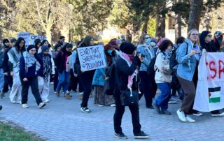 A group of people, including members from pro Palestinian clubs, participate in an outdoor protest along a brick pathway, holding signs like "END THAT SHEIKH JARRAH." Many wear keffiyehs as they raise their voices for Palestine, with trees standing tall in the background.