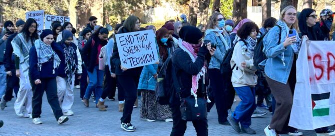 A group of people, including members from pro Palestinian clubs, participate in an outdoor protest along a brick pathway, holding signs like "END THAT SHEIKH JARRAH." Many wear keffiyehs as they raise their voices for Palestine, with trees standing tall in the background.