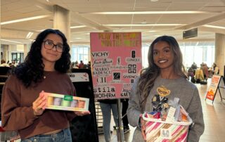 Two people stand indoors, holding gift baskets in front of a raffle table adorned with a poster showcasing various brand logos. In the well-lit space bustling with activity, members of the Pre-Law club engage in an exciting charity event.