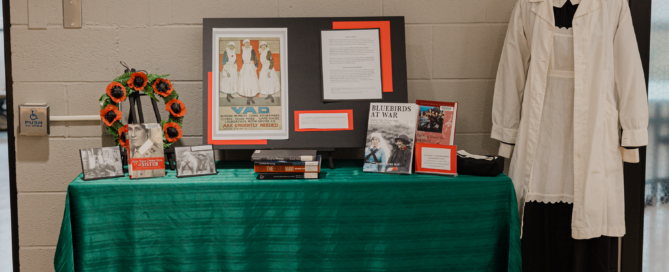 A table draped in green at St. Jerome’s University showcases a framed poster, books, photos, and wartime memorabilia. Highlighting the role of wartime nurses, a mannequin dons a vintage nurse uniform. A wreath for Remembrance Day and informational text complete the display.