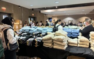 People are gathered around tables with stacks of folded clothing, such as hoodies and sweatshirts, for sale in a well-lit indoor W Store pop-up event. Some customers are making selections, while others browse or chat.