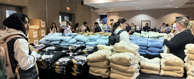 People are gathered around tables with stacks of folded clothing, such as hoodies and sweatshirts, for sale in a well-lit indoor W Store pop-up event. Some customers are making selections, while others browse or chat.