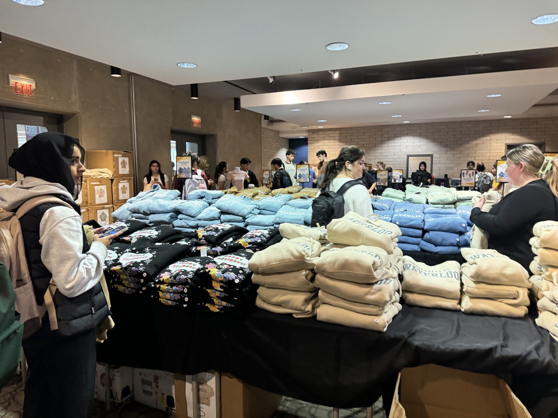 People are gathered around tables with stacks of folded clothing, such as hoodies and sweatshirts, for sale in a well-lit indoor W Store pop-up event. Some customers are making selections, while others browse or chat.