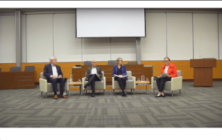 A panel of four people seated in an auditorium, transitioning smoothly into the discussion. They sit in armchairs with microphones and water glasses on side tables. Behind them is a large blank screen, with a lectern to the right. The room is empty except for the panelists from UW addressing the hiring freeze.