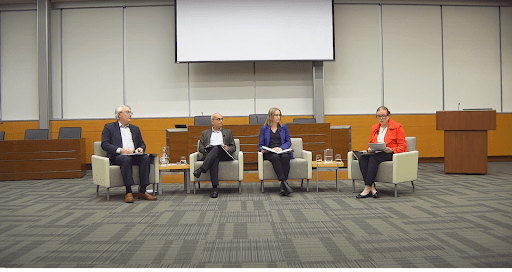 A panel of four people seated in an auditorium, transitioning smoothly into the discussion. They sit in armchairs with microphones and water glasses on side tables. Behind them is a large blank screen, with a lectern to the right. The room is empty except for the panelists from UW addressing the hiring freeze.