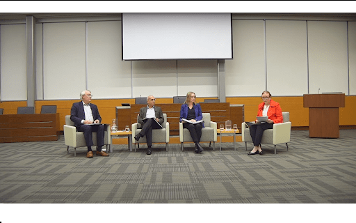 A panel of four people seated in an auditorium, transitioning smoothly into the discussion. They sit in armchairs with microphones and water glasses on side tables. Behind them is a large blank screen, with a lectern to the right. The room is empty except for the panelists from UW addressing the hiring freeze.