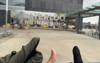 Three hands showing thumbs up in front of the University of Waterloo Engineering building, just a 75 km trek from Toronto. The building boasts reflective glass walls and a modern design. Bicycles are parked to the right. One hand sports a green glove, another a black glove, while the third is bare.