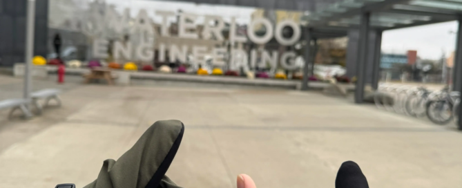 Three hands showing thumbs up in front of the University of Waterloo Engineering building, just a 75 km trek from Toronto. The building boasts reflective glass walls and a modern design. Bicycles are parked to the right. One hand sports a green glove, another a black glove, while the third is bare.