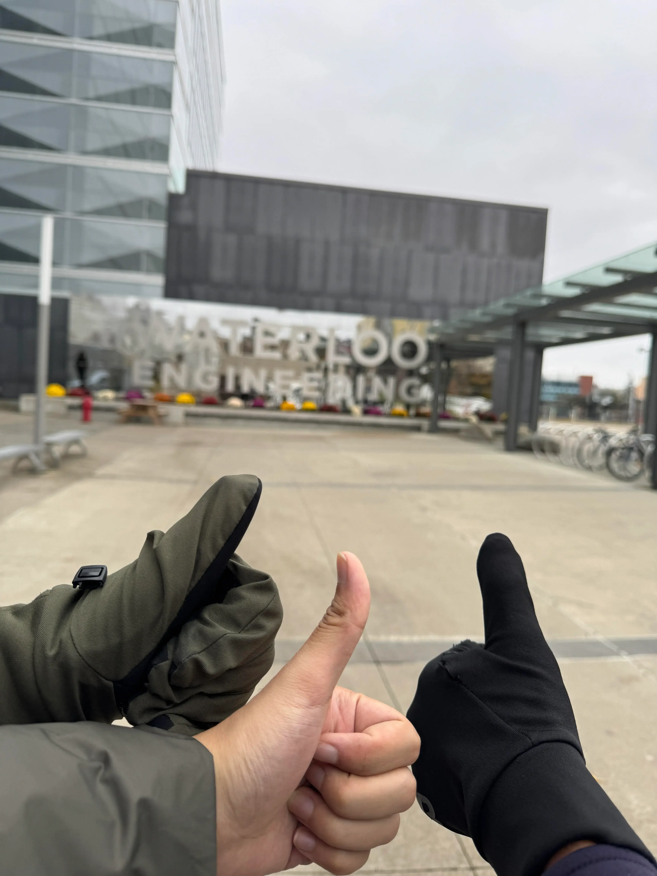 Three hands showing thumbs up in front of the University of Waterloo Engineering building, just a 75 km trek from Toronto. The building boasts reflective glass walls and a modern design. Bicycles are parked to the right. One hand sports a green glove, another a black glove, while the third is bare.