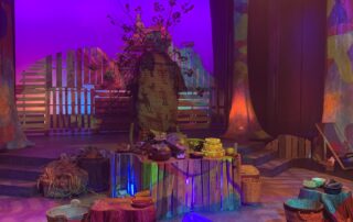 A colorful stage set with wooden props, baskets, and tree stumps under warm lighting evokes the mystical tale of Th'owxiya. A backdrop of purple and blue hues creates an enchanting ambience, with a central structure resembling a large, decorated egg surrounded by branches.