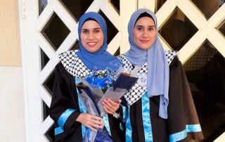 Two women in graduation gowns and hijabs stand in front of a white door. One holds a bouquet and a diploma. Both wear blue and black outfits with matching scarves and look towards the camera, smiling.