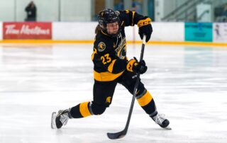 A women's hockey player in a black and yellow uniform, number 23, skates swiftly across the ice under the banners of UW. As she holds her stick ready to shoot the puck, the arena's empty spectator area echoes her commitment to excellence fostered by the national coaching program.